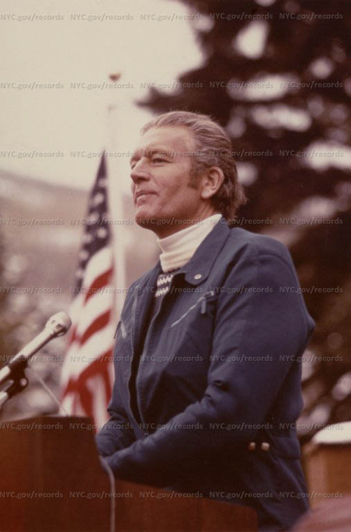 John Lindsay standing at outdoor lectern: American flag in background. John Lindsay wearing turtle neck shirt.